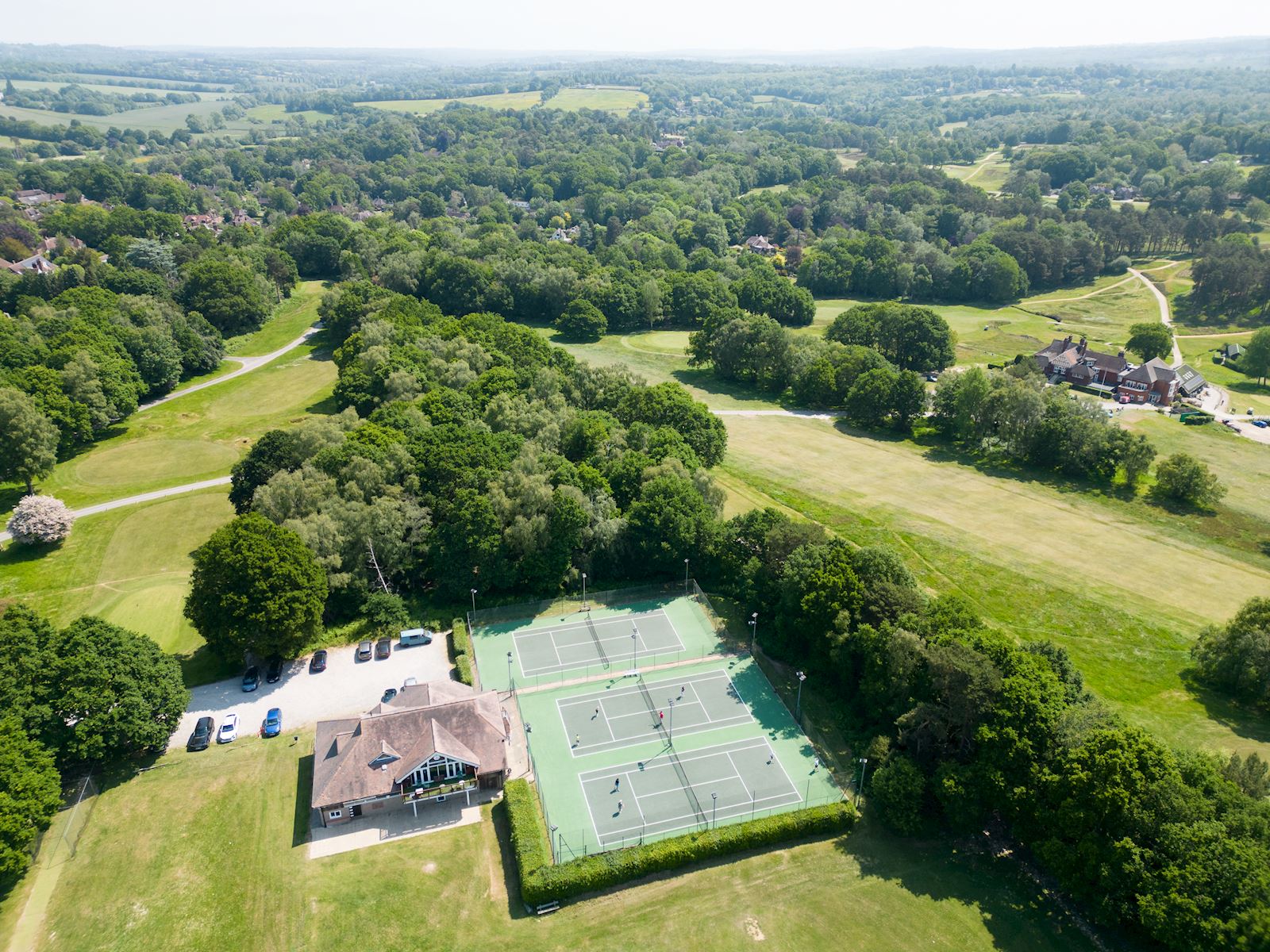Aerial view of Forest Row Lawn Tennis Club, with players on the three all-weather courts beside the clubhouse, surrounded by trees and open countryside